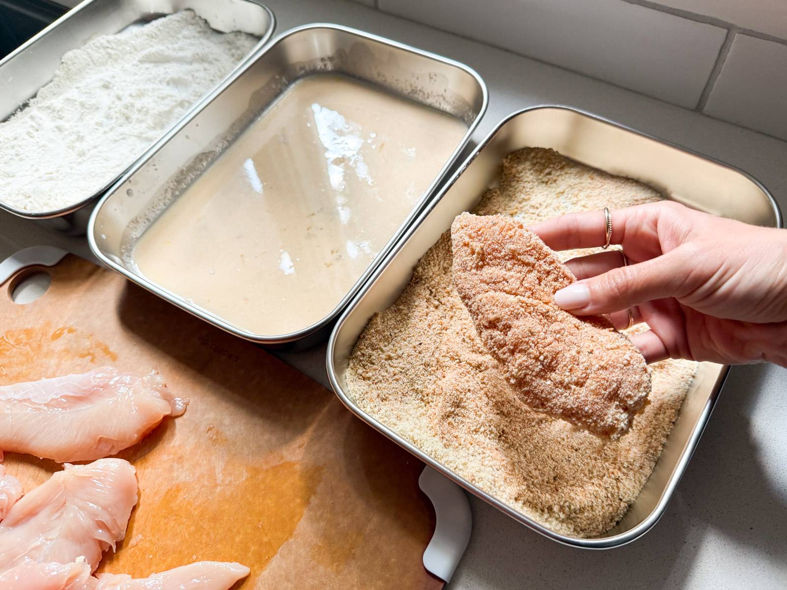 Three containers filled with flour, egg wash, and breadcrumbs with chicken tenderloins.